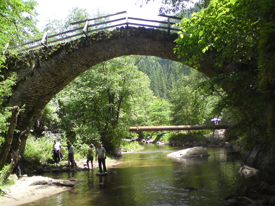 Stone mystical bridge over a calm river surrounded by lush green trees with people exploring the waters below.