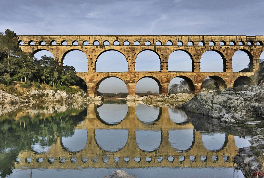 Ancient mystical stone bridge with multiple arches reflecting in calm river water surrounded by nature and rocky terrain.