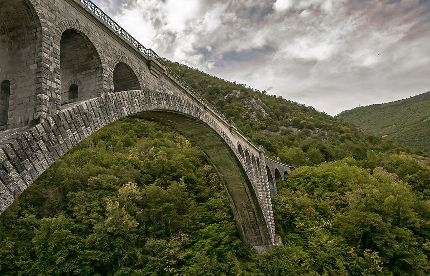 Stone arch bridge towering over dense green forest beneath a cloudy sky, showcasing a mystical bridge in nature.