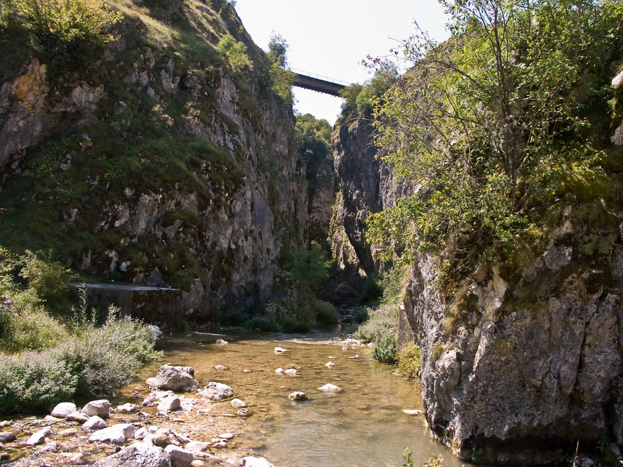 A mystical bridge spanning a deep rocky gorge over a calm river surrounded by lush greenery.