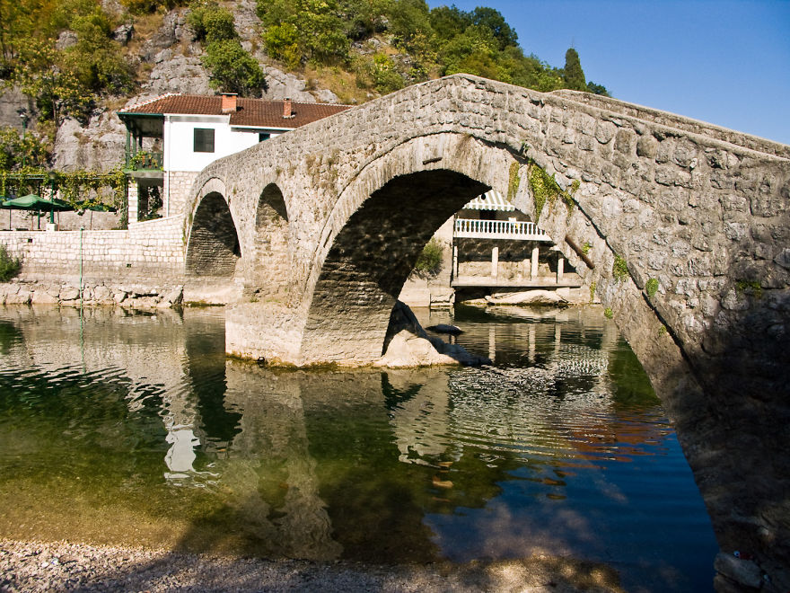 Ancient mystical stone bridge over calm water surrounded by greenery and rocky hills on a sunny day