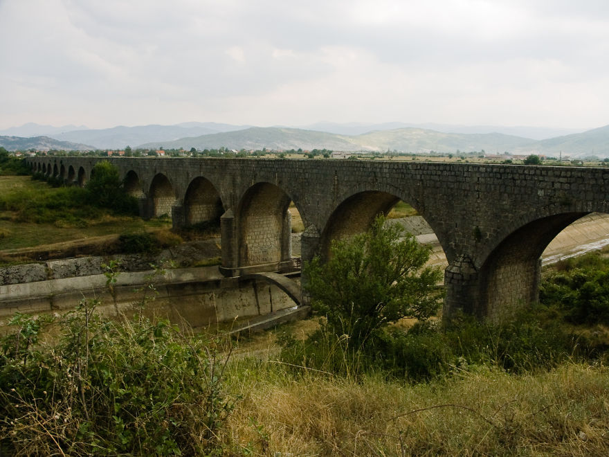 Ancient mystical stone bridge with multiple arches spanning a dry riverbed, surrounded by greenery and distant mountains.