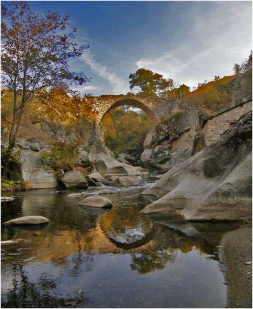 Ancient mystical stone bridge arching over a calm river surrounded by autumn trees and large rocks.