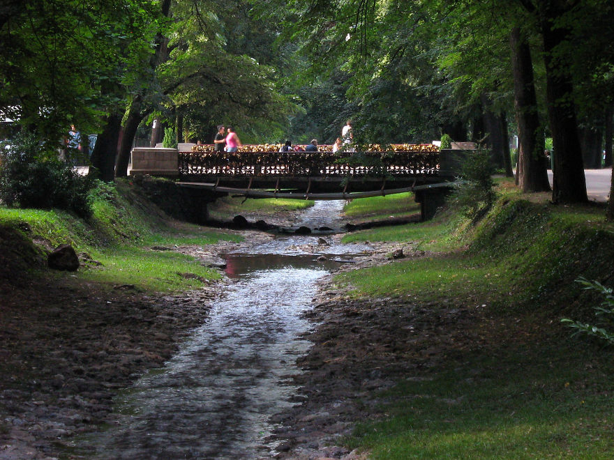 Mystical bridge over a calm stream surrounded by lush trees with people walking on the bridge in a serene park setting