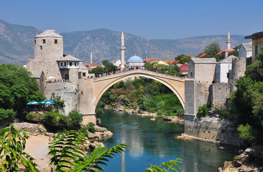 Stone bridge arching over a river surrounded by historic buildings and lush greenery in a mystical bridges landscape.
