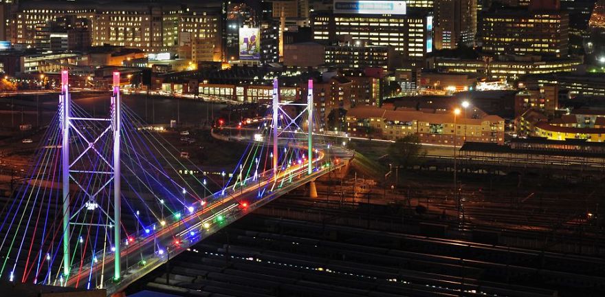Colorfully illuminated mystical bridge lit up at night, stretching over railway tracks with a city skyline in the background.