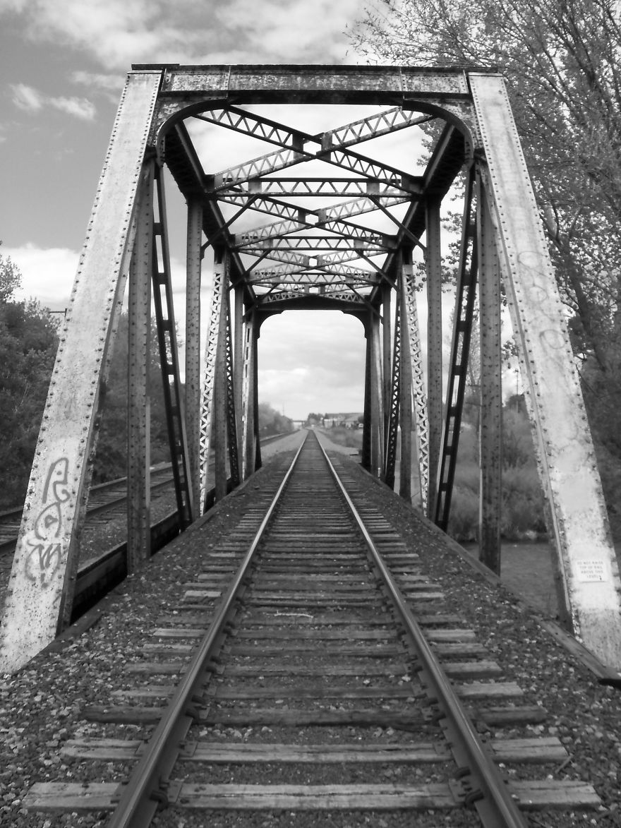Rustic steel railway bridge with tracks stretching into the distance, surrounded by trees and cloudy sky in black and white.