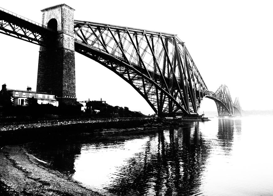 Black and white image of a mystical bridge with intricate ironwork arching over calm water, reflecting the structure below.