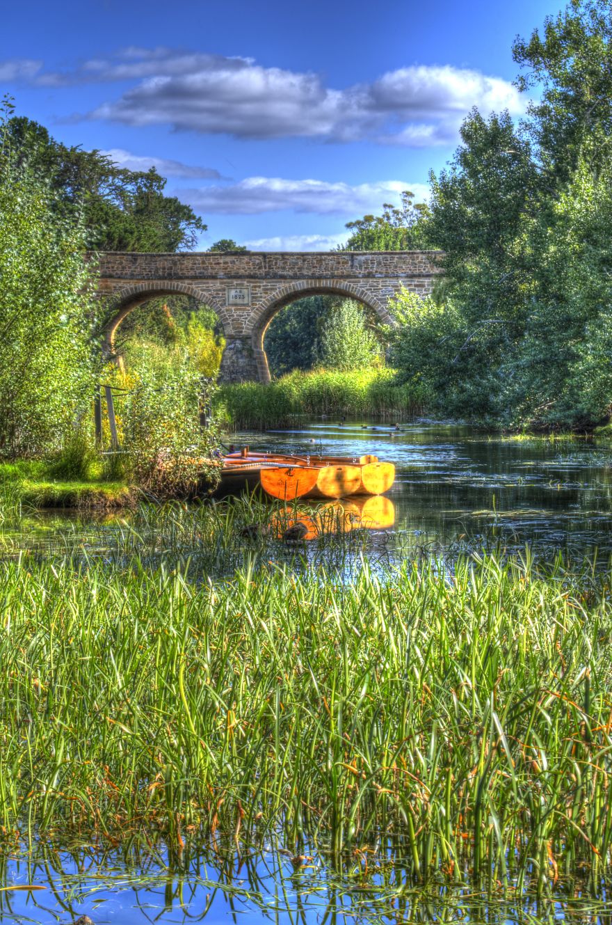 Stone bridge with arches over a calm river surrounded by lush greenery and a wooden boat reflecting on the water.