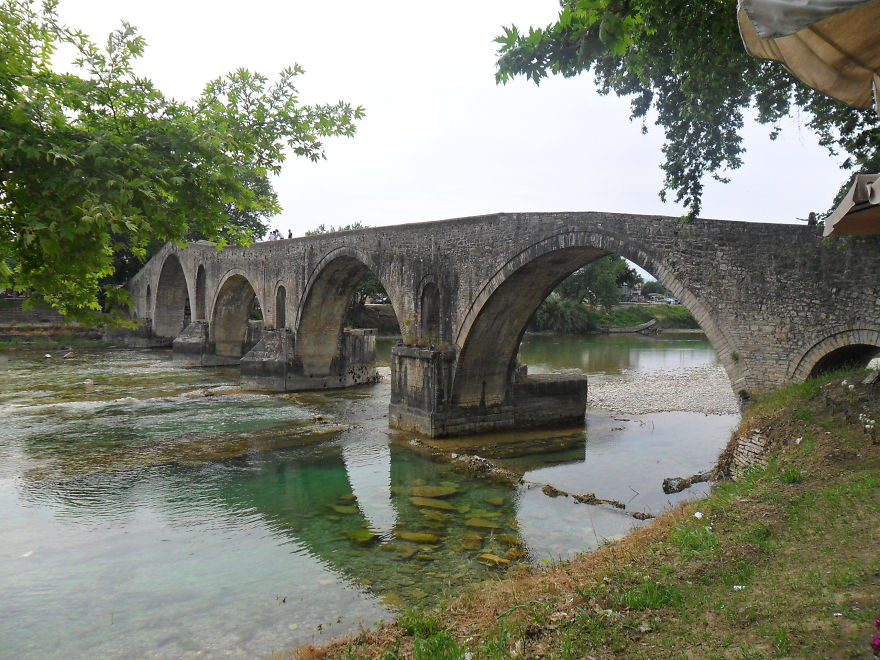 Ancient mystical stone bridge with arches crossing clear river water surrounded by trees and greenery.