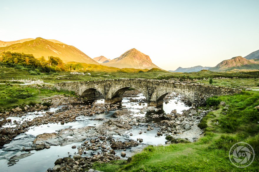 Stone arch mystical bridge over a rocky river with mountains in the background during golden hour light
