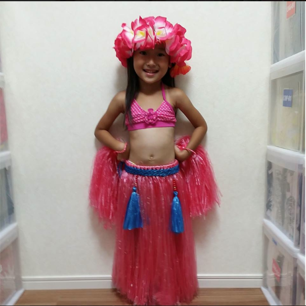 Young girl smiling in a colorful children's Halloween costume with a pink flower headpiece and grass skirt indoors.