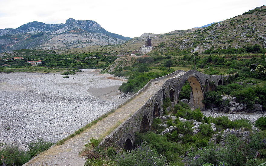 Ancient mystical stone bridge with multiple arches crossing a dry riverbed surrounded by mountains and greenery.