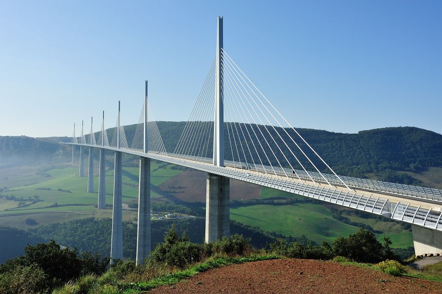 Modern mystical bridge spanning a deep valley with mountains and greenery under a clear blue sky.