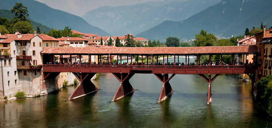 Covered wooden bridge over calm river with historic buildings and mountains in the background, showcasing mystical bridges.
