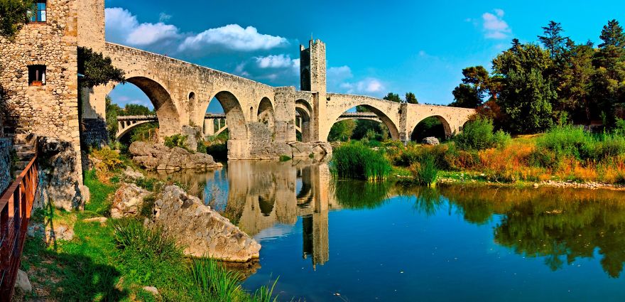 Ancient mystical stone bridge with arches reflecting over a calm river surrounded by greenery under a blue sky.
