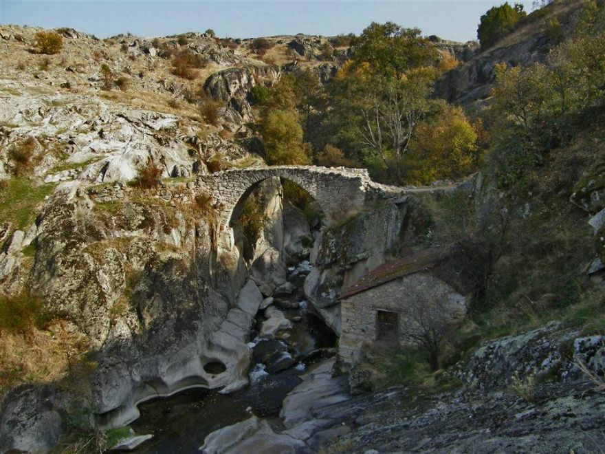 Ancient mystical stone bridge arching over rocky river gorge surrounded by rugged terrain and sparse trees.