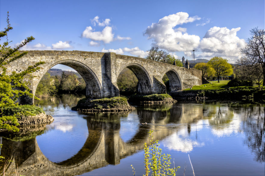 Stone arch mystical bridge over calm river surrounded by green trees under a bright blue sky with clouds.
