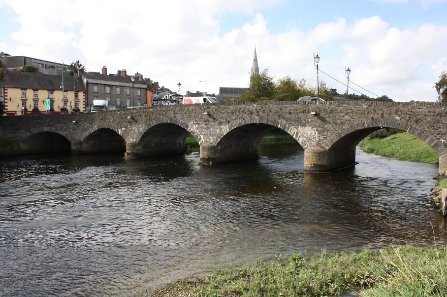 Stone mystical bridge with multiple arches spanning a river near a small town under a partly cloudy sky.