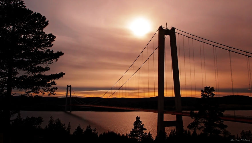 Suspension bridge at sunset over calm water surrounded by trees, showcasing a mystical and serene atmosphere.