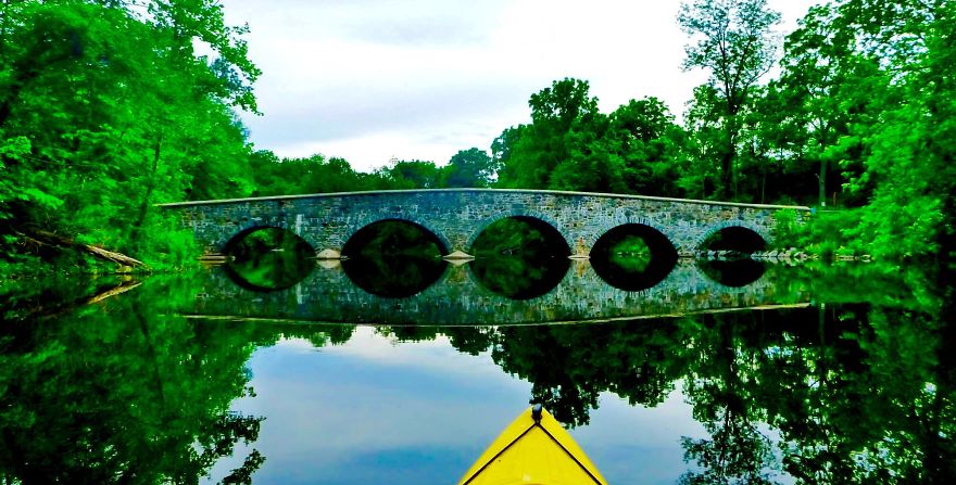 Stone mystical bridge over calm water surrounded by lush green trees, reflecting a peaceful and otherworldly scene.