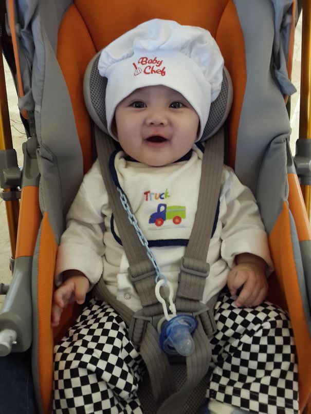 Baby in children's Halloween costume ideas dressed as a baby chef sitting in an orange stroller harnessed securely.