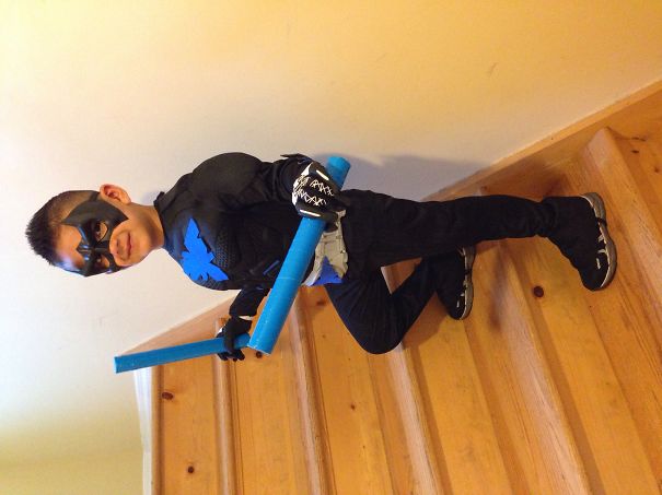 Young boy posing on wooden stairs in a black and blue superhero costume holding blue foam weapons for Halloween costume ideas.