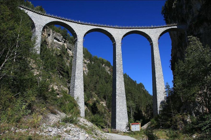 Tall mystical stone bridge with large arches spanning a forested valley under a clear blue sky.