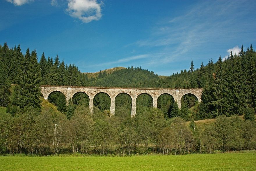 Stone arch mystical bridge surrounded by forest under a blue sky, blending nature and ancient architecture in a serene landscape.