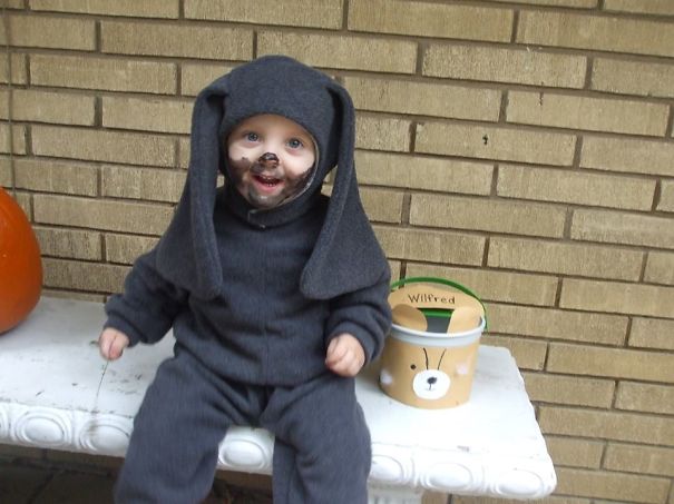 Toddler in a black dog costume with face paint, sitting on a bench next to a Halloween bucket, children’s Halloween costume ideas.