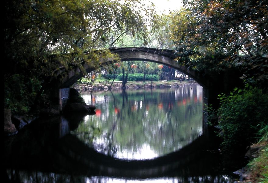 Stone arch mystical bridge over calm water surrounded by lush trees, reflecting a serene and otherworldly atmosphere.