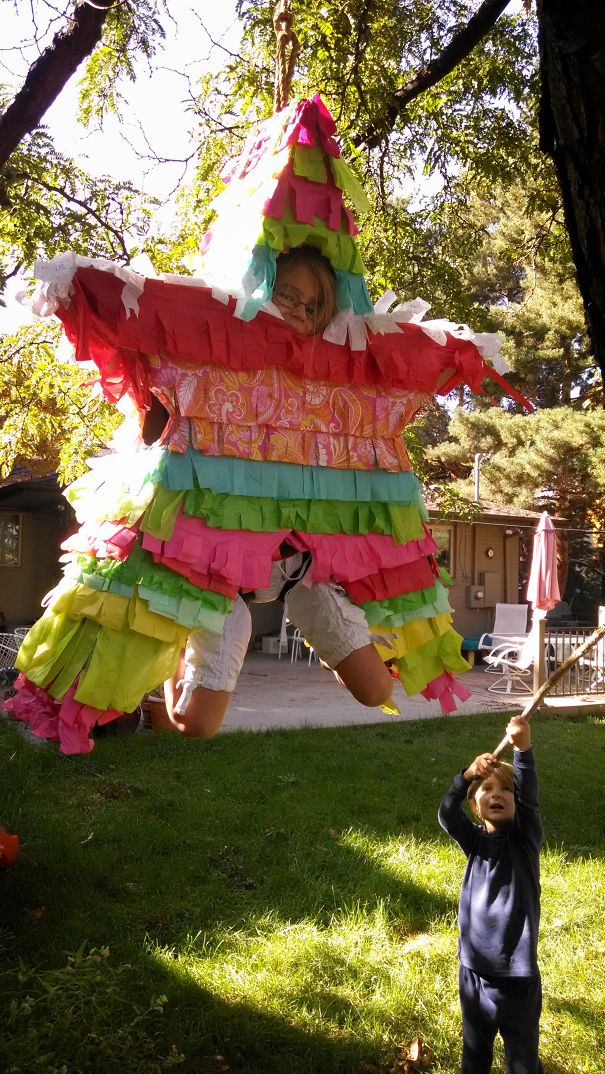 Child in colorful pinata costume hanging from tree with another child holding stick in backyard setting, children's Halloween costume ideas.
