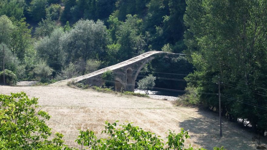 Ancient mystical stone bridge arching over a river surrounded by lush green trees and a clearing in the countryside.