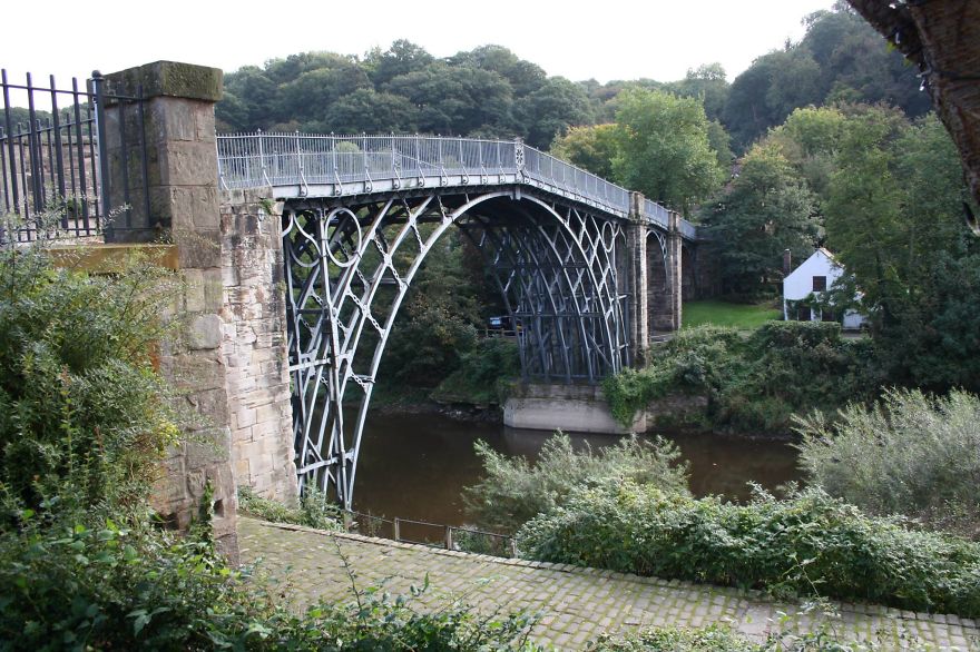 Iron bridge arching over calm river surrounded by lush greenery, showcasing mystical bridges in a serene natural setting.