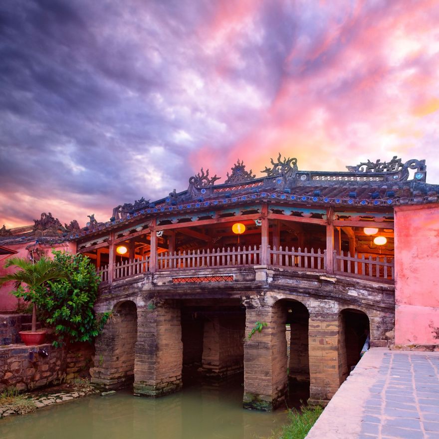 Ancient mystical bridge at sunset with intricate woodwork and stone pillars over calm water in a vibrant sky setting