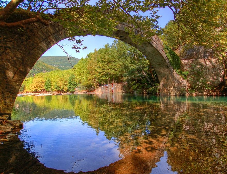 Ancient mystical stone bridge arching over calm river surrounded by lush green trees and clear reflections.