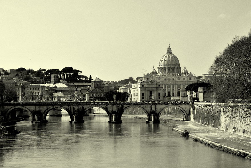 Mystical bridge over calm river with historic buildings and a large dome in the background during daylight.
