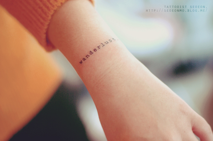 A minimalistic tattoo by Seoeon featuring the word "wanderlust" in simple lettering on a person's wrist. A minimalistic tattoo by Seoeon featuring the word "wanderlust" in simple lettering on a person's wrist.