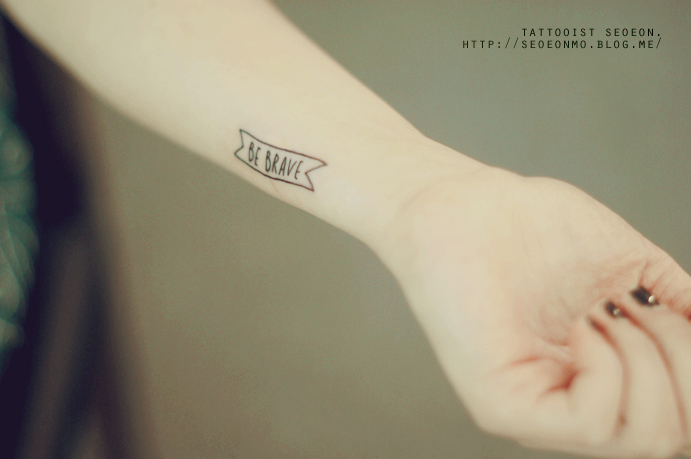 Minimalistic tattoo by Seoeon on a wrist, featuring a small banner with the text "Be Brave. Minimalistic tattoo by Seoeon on a wrist, featuring a small banner with the text "Be Brave.