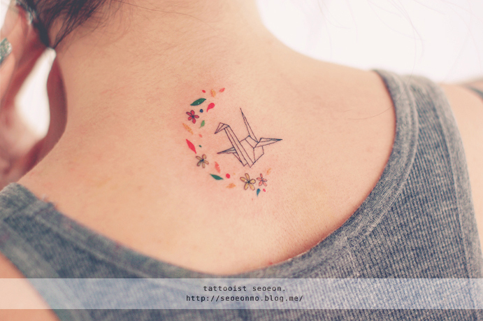Minimalistic tattoo by Seoeon on a person's neck, featuring a delicate origami crane surrounded by colorful floral accents.