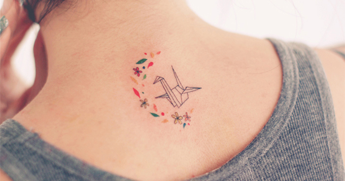 Minimalistic tattoo of a delicate origami crane surrounded by small colorful flowers on the upper back of a person