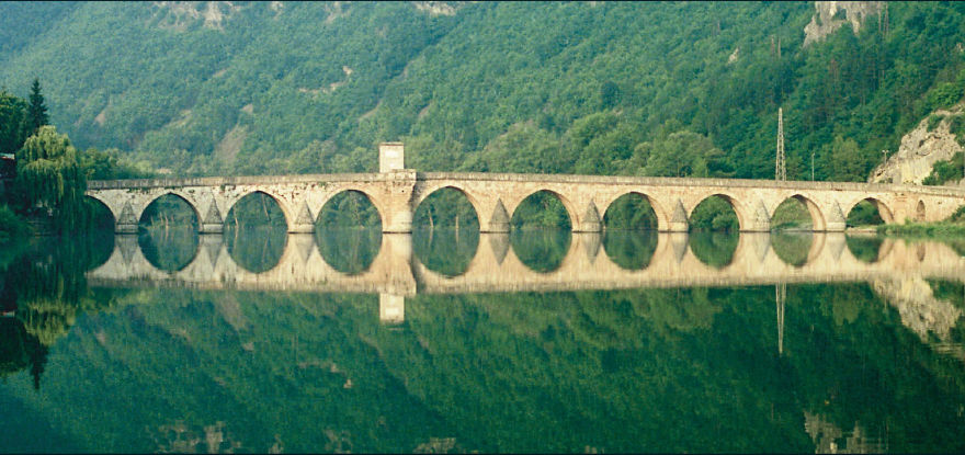 Ancient mystical bridge with multiple arches reflecting on calm water surrounded by lush green forest and hills.