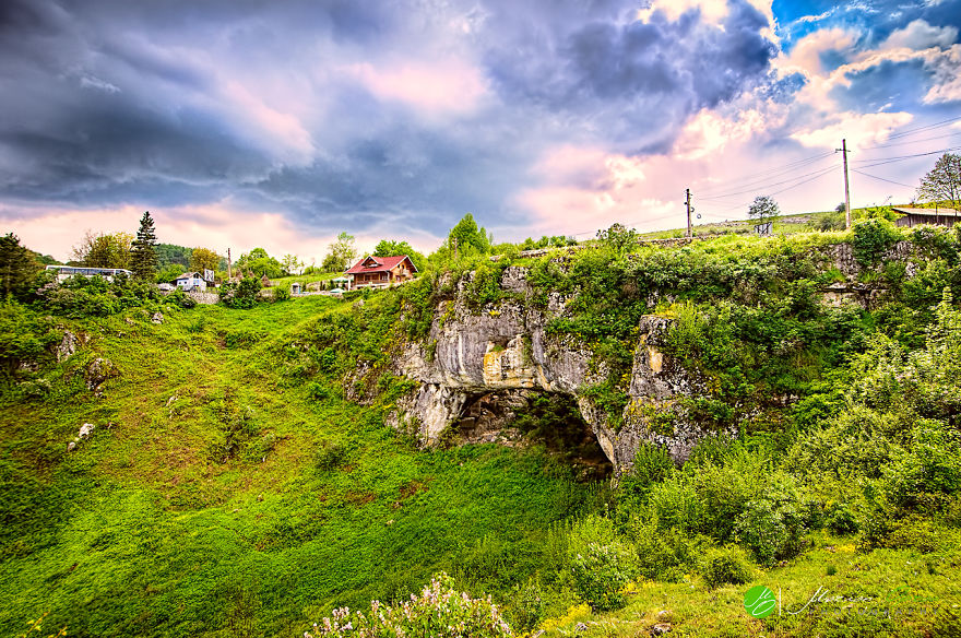 Lush green landscape with a natural rock bridge under a dramatic cloudy sky in a mystical setting.