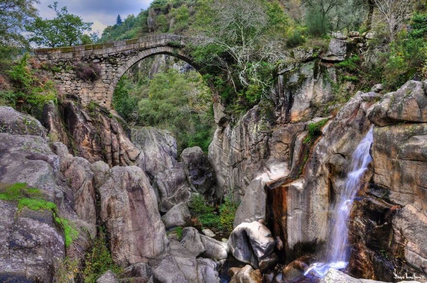 Ancient mystical stone bridge arching over rocky terrain with lush greenery and a cascading waterfall nearby in a forest setting.