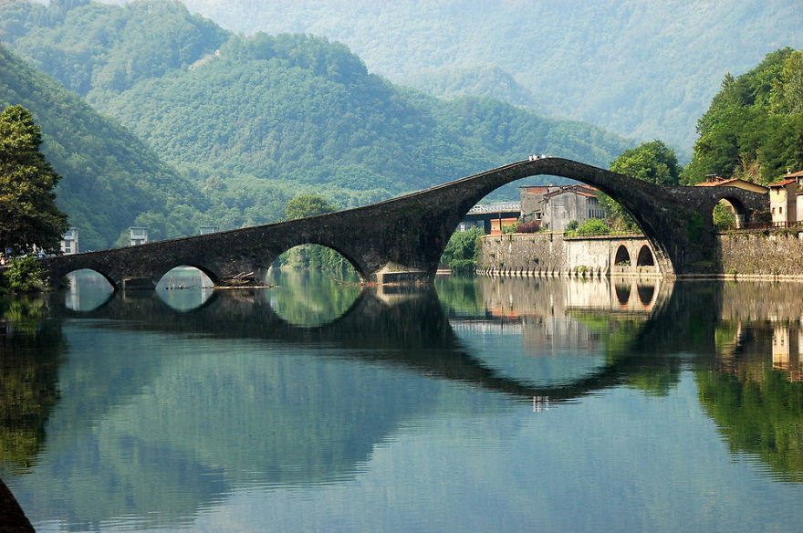 Ancient mystical stone bridge with multiple arches reflected in calm river surrounded by lush green hills and small village.
