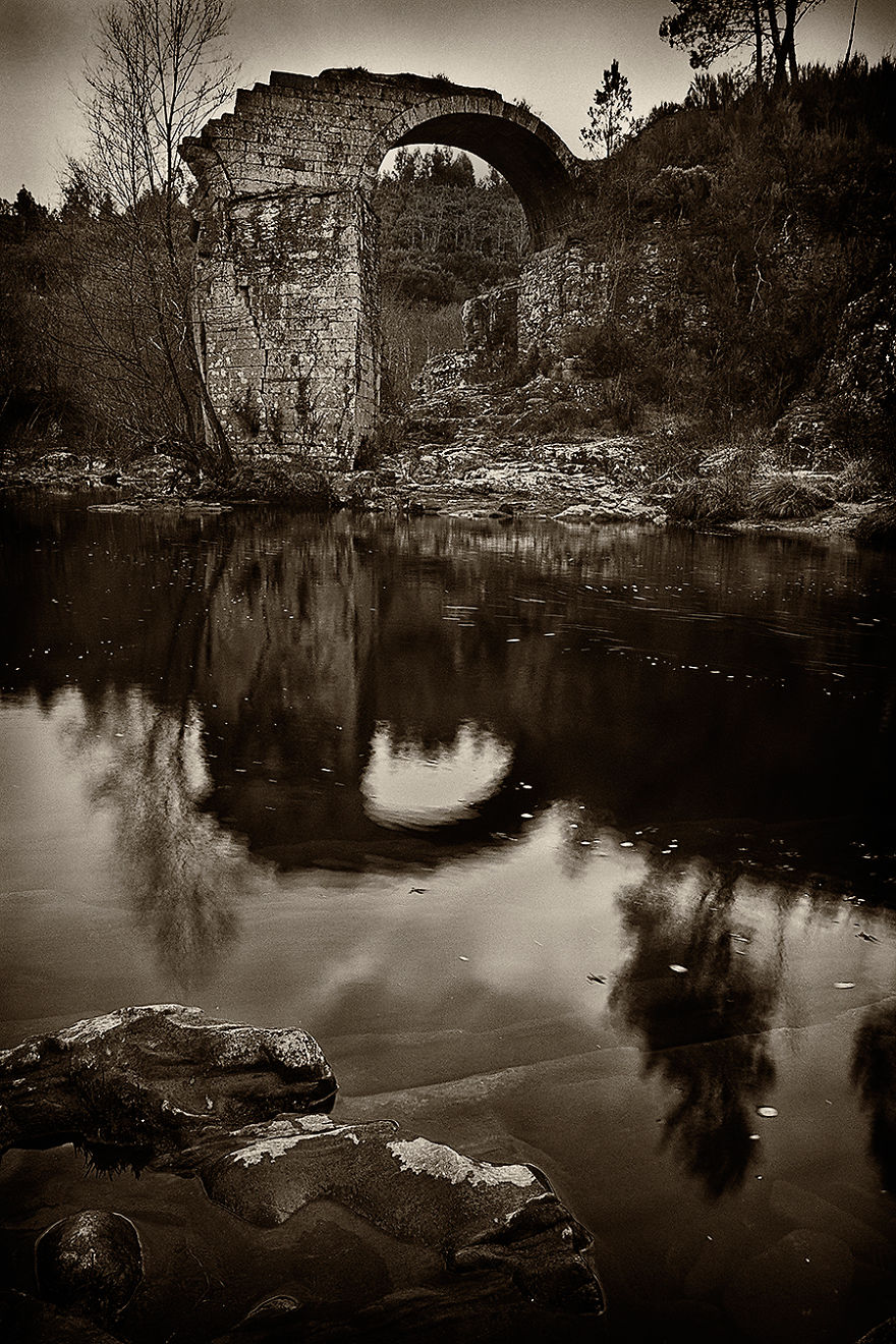 Ancient mystical stone bridge arch reflecting in calm river water surrounded by forest and rocky terrain at dusk.