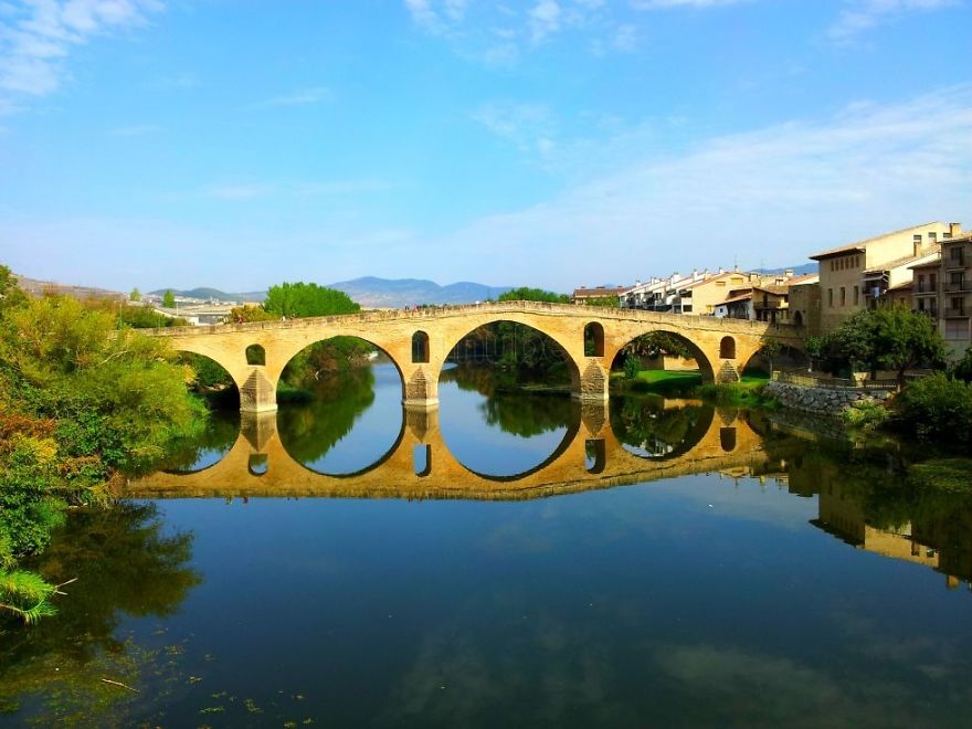 Ancient mystical stone bridge with arches reflected on calm river water under a bright blue sky in a peaceful landscape.