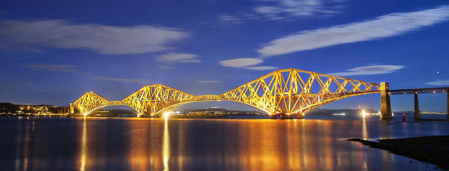 Illuminated mystical bridge at night reflecting on calm water under a clear sky, evoking otherworldly beauty and enchantment.