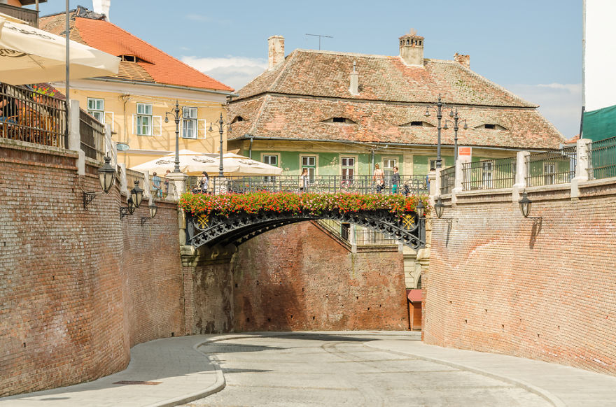 Iron bridge adorned with flowers over a narrow street, surrounded by historic buildings and brick walls in a mystical setting.