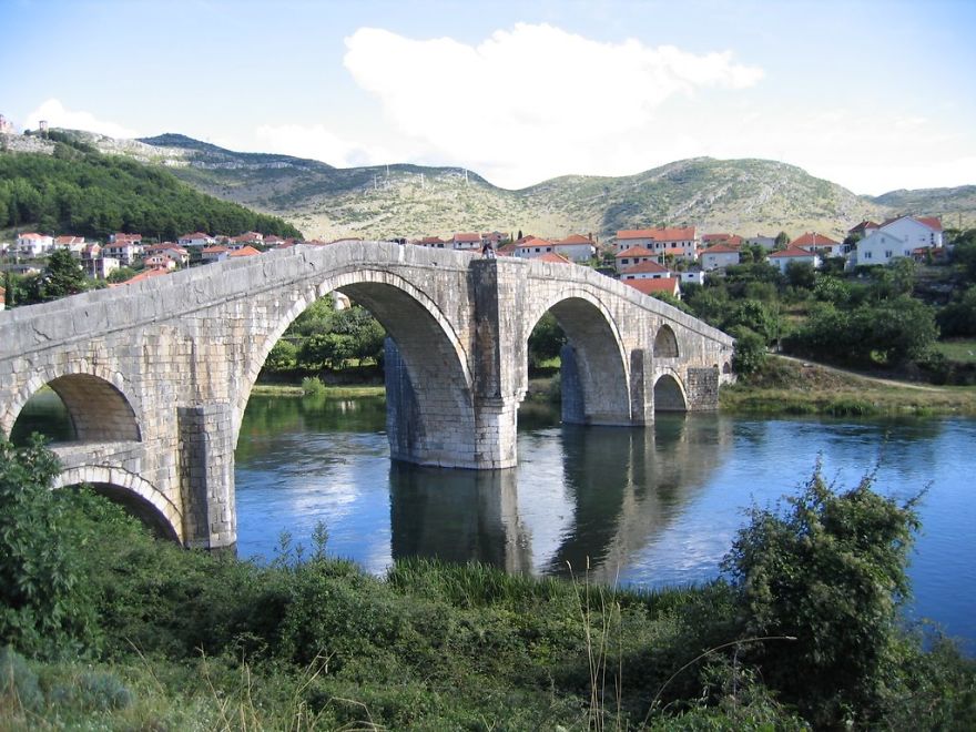Ancient mystical stone arch bridge over calm river surrounded by greenery and village houses under blue sky